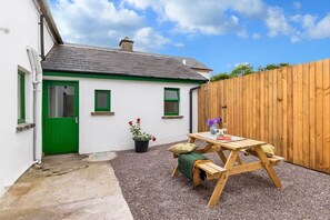 A white house with a green door, a wooden fence, and a gravel patio with a wooden table and chairs. - Kenmare Pier Cottage 
Cosy sea side home . (Kenmare)