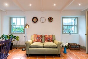 Un salón con un sofá, dos relojes de pared, una lámpara de pie, una mesita con un florero y un ventanal con vistas.