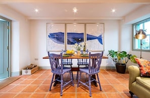 A dining area with a table set for four, chairs, a wall-mounted artwork, a potted plant, and a window with natural light. - Kenmare Pier Cottage 
Cosy sea side home . (Kenmare)