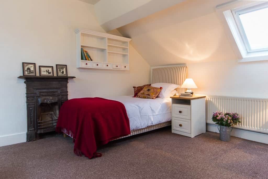 A bedroom with a bed, a nightstand, a red throw blanket, a fireplace, a radiator, a shelf with books, and a window.