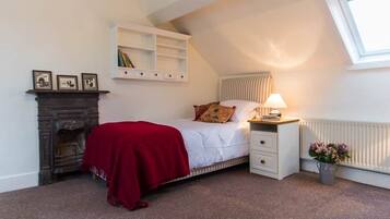 A bedroom with a bed, a nightstand, a red throw blanket, a fireplace, a radiator, a shelf with books, and a window.