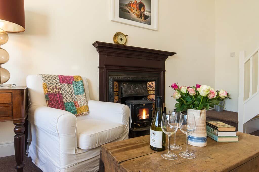 Un salon chaleureux avec une cheminée, un canapé blanc, une table basse en bois sur laquelle se trouvent une bouteille de vin et des verres, et un vase rempli de fleurs.