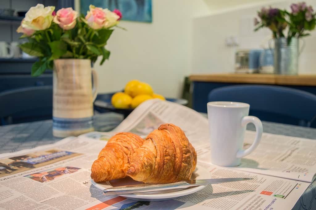 Een croissant op een bord, een witte mok en een krant op een tafel, met een bloevas op de achtergrond.