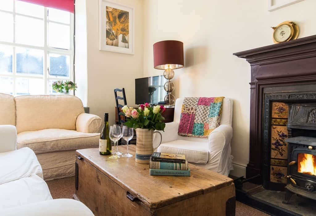 A cozy living room with a fireplace, a sofa, a coffee table with books and glasses, and a lamp.