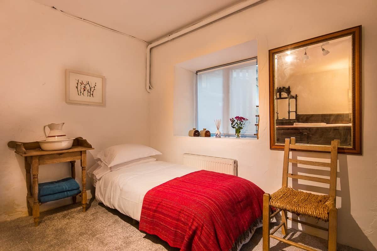 Un dormitorio con una cama, una silla, una mesa con un cuenco, un radiador, una ventana con flores y un espejo enmarcado.
