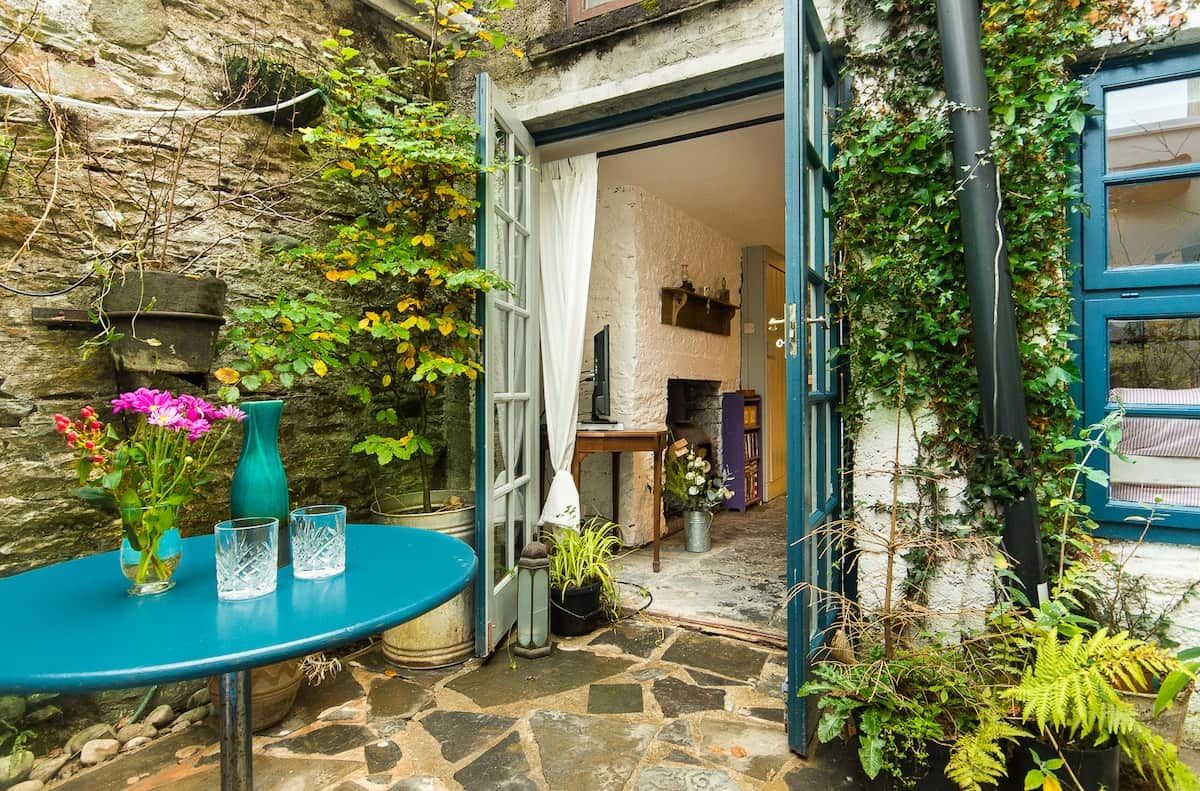 Un espacio exterior con suelo de piedra, una mesa azul con un florero y vasos, y una puerta que da a una habitación con pared blanca y ventana azul.