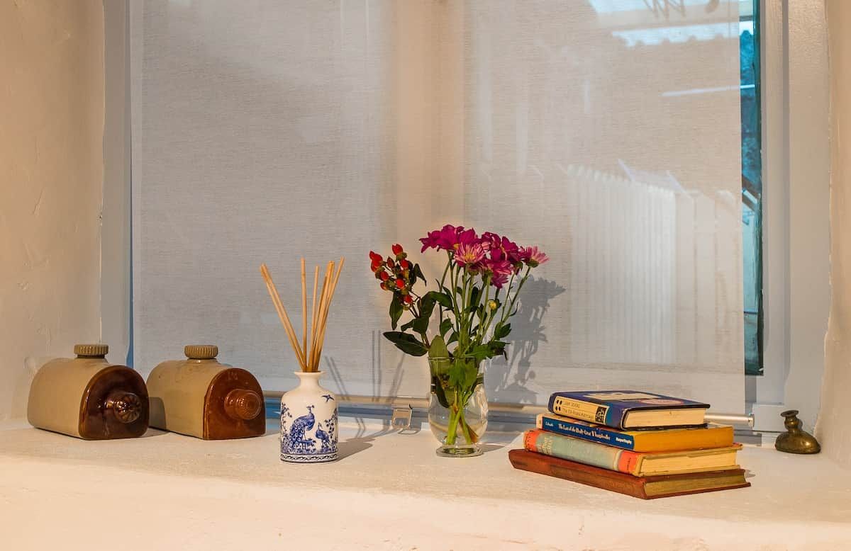 Ein Fensterbrett mit einer Blumenvase, zwei dekorativen Behältern und einem Stapel Bücher.