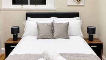 A neatly made bed with a quilted cover, two bedside tables with lamps, a window with a blind, and a wall-mounted artwork.