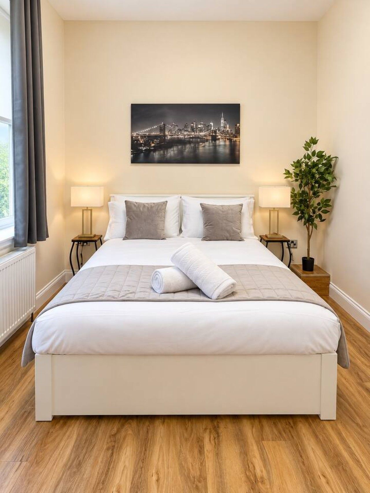 Ein Hotelzimmer mit einem großen Bett, zwei Nachttischen mit Lampen, Blick auf eine Stadtlandschaft und einer Zimmerpflanze.