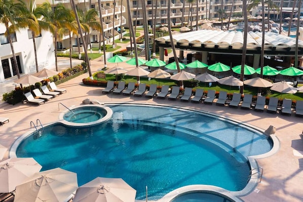 A resort pool area with multiple lounge chairs and umbrellas, surrounded by palm trees and a building with balconies.