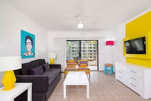 A modern living room with a sofa, a coffee table, a TV on a yellow stand, and a balcony with a view of buildings.
