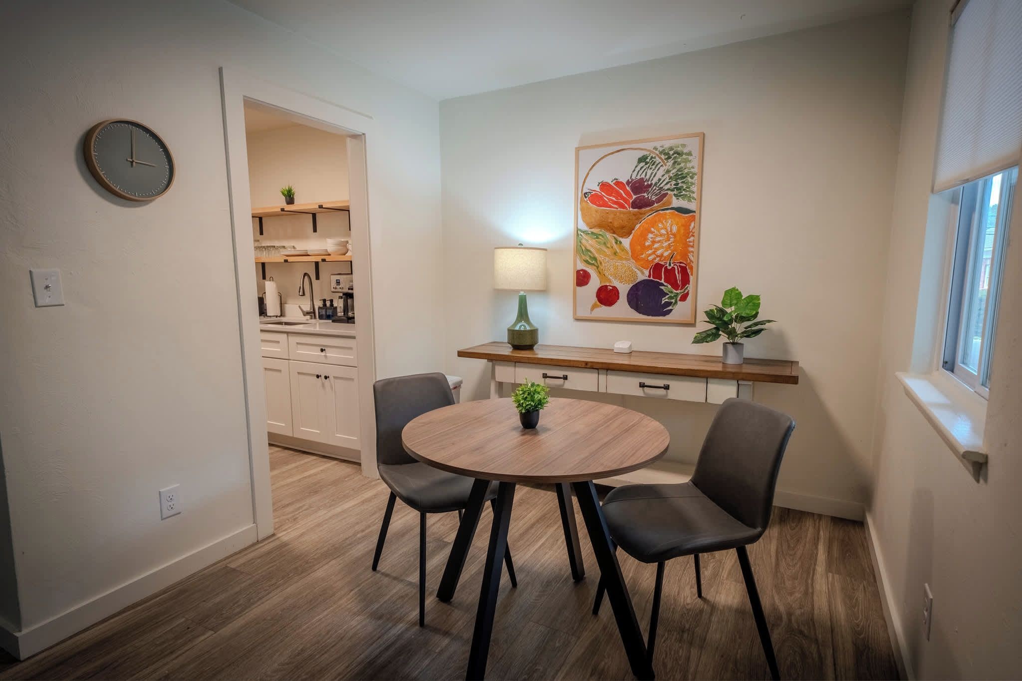 Un petit coin repas comprenant une table ronde, deux chaises, une horloge murale et un tableau représentant des fruits.