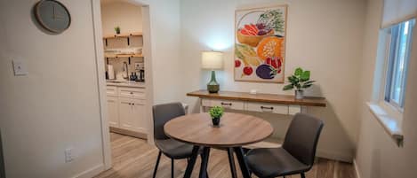 A compact dining area with a round table, two chairs, a wall clock, and a fruit painting.