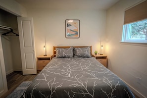 Ein Schlafzimmer mit einem Bett, zwei Nachttischen, einer Tür, einem Fenster mit Jalousien und einem an der Wand hängenden abstrakten Gemälde.
