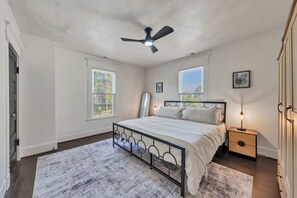 A bedroom with a bed, a ceiling fan, two windows, a nightstand, and a wall-mounted lamp. - Stay Roanoke | Patterson Pad | Roomy 3BR in Town (Roanoke)