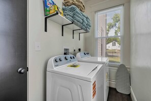 A laundry room with two front-loading washers and dryers, a shelf with cleaning supplies, and a window with blinds. - Stay Roanoke | Patterson Pad | Roomy 3BR in Town (Roanoke)