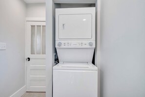 A compact, white laundry room with a front-loading washer and dryer, a door with frosted glass panels, and a tiled floor. - Stay Roanoke | 4BR near River, Greenway, Families (Roanoke)
