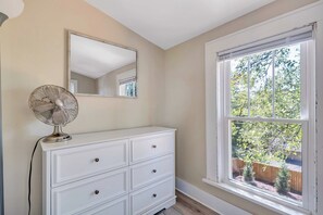 Een kamer met een witte ladekast, een ventilator, een spiegel en een raam met uitzicht op bomen.