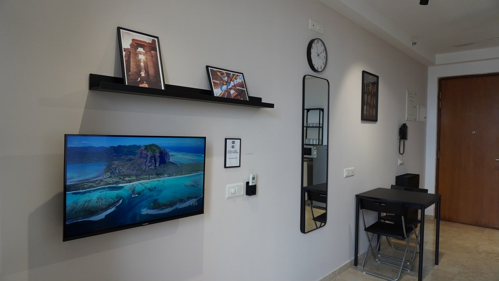 Een kamer met een wandtelevisie, een bureau met een stoel, een spiegel en een klok.