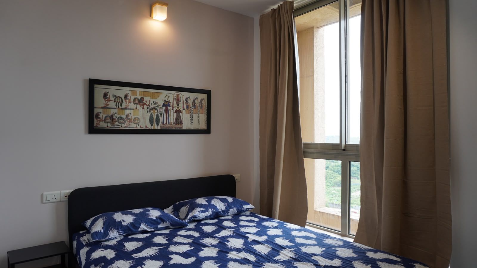 A bedroom with a bed, a nightstand, a wall-mounted light, a window with curtains, and a framed picture on the wall.