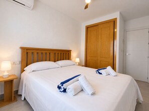 A hotel room with a wooden headboard, two white pillows, a bed with white linens, a nightstand with a lamp, and a wooden wardrobe. - Jardin del Mar House Torrevieja (Torrevieja)