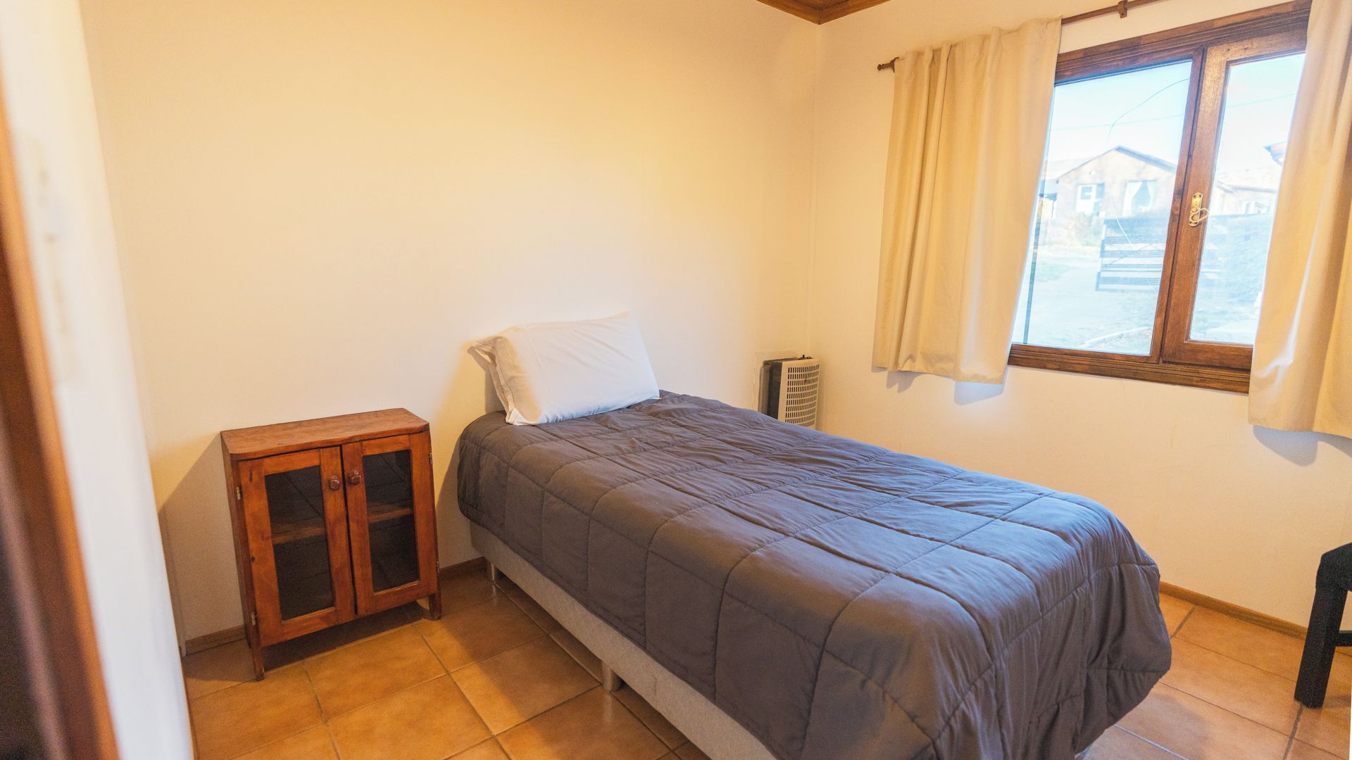 Un lit simple avec une tête de lit, une petite armoire en bois, un radiateur et une fenêtre avec des rideaux.