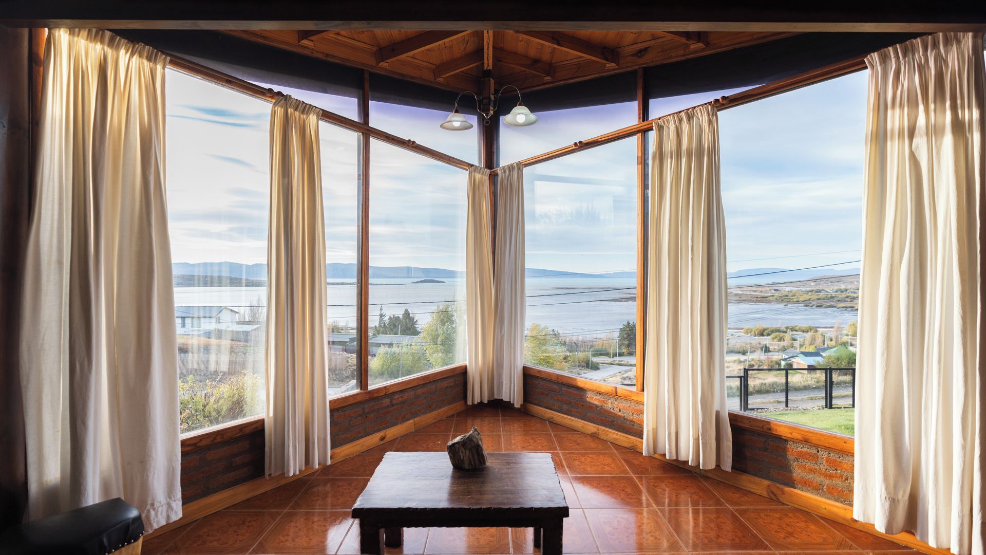 Quarto de canto com amplas janelas, uma mesa de madeira e vista para a paisagem.