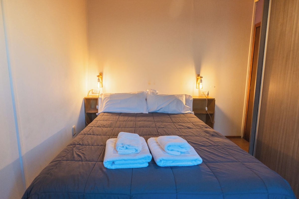 A narrow hotel room with a single bed, two bedside lamps, and a wooden door.
