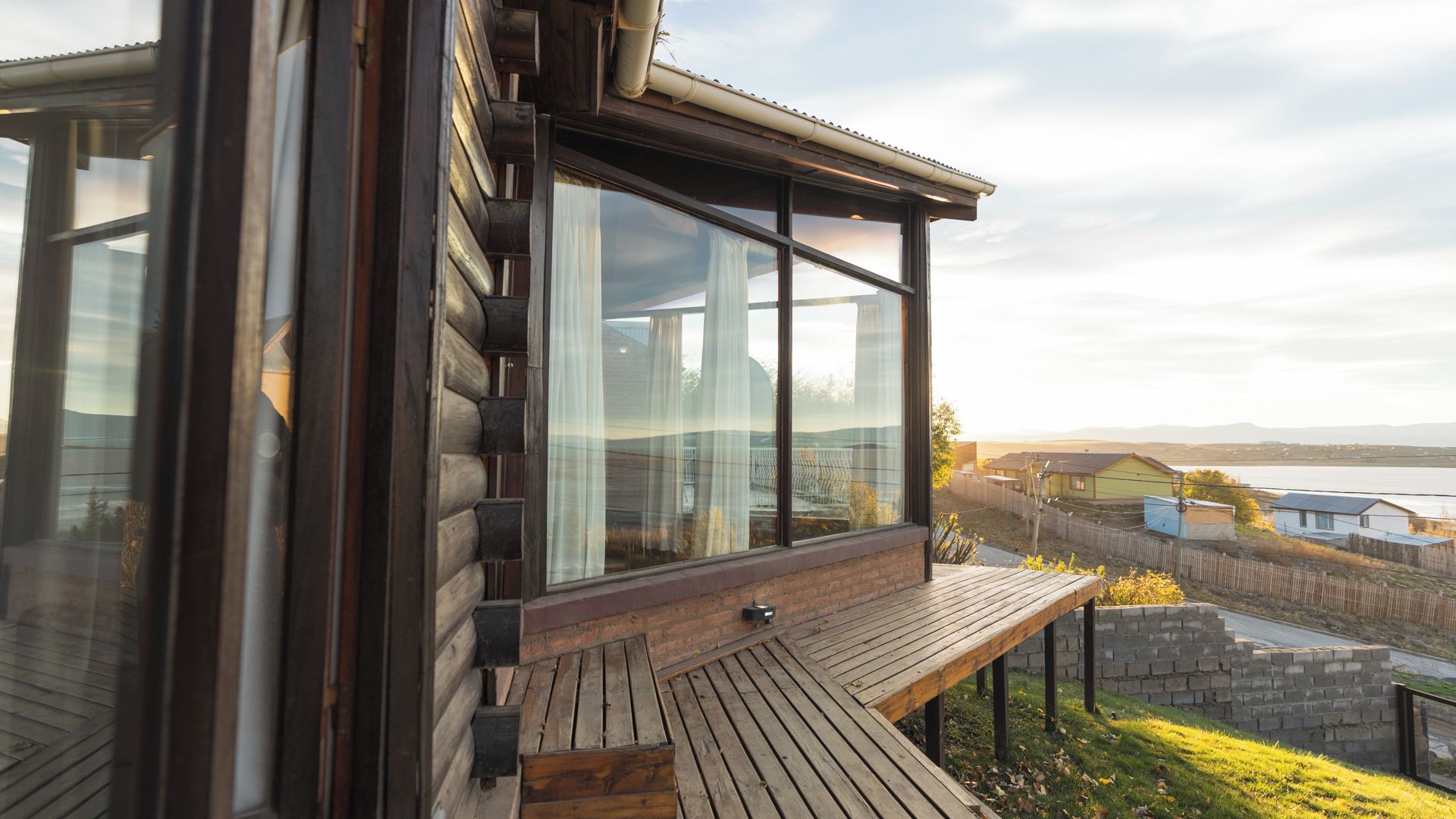 Une cabane en rondins moderne, dotée de grandes fenêtres, d’une terrasse en bois et offrant une vue sur un lac et des maisons situées au loin.