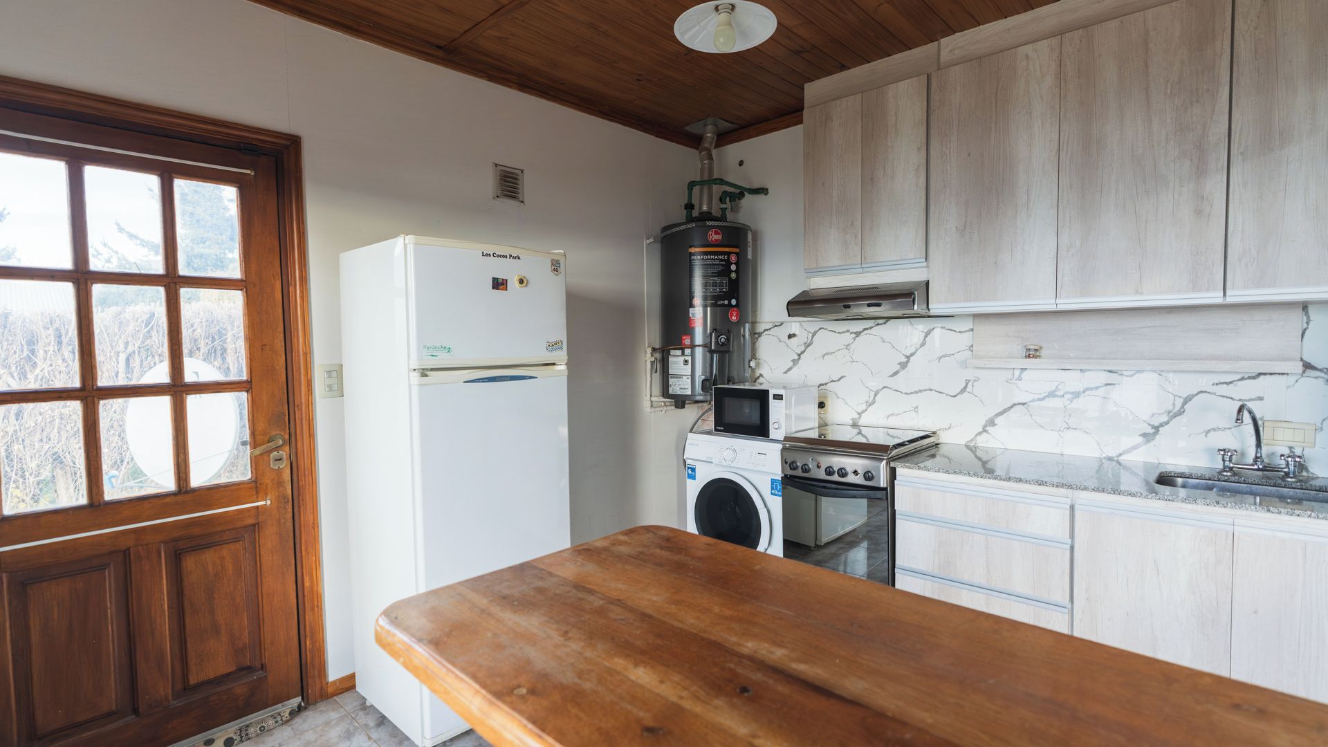 A compact kitchen with a wooden table, a refrigerator, a washing machine, a microwave, and a stove.