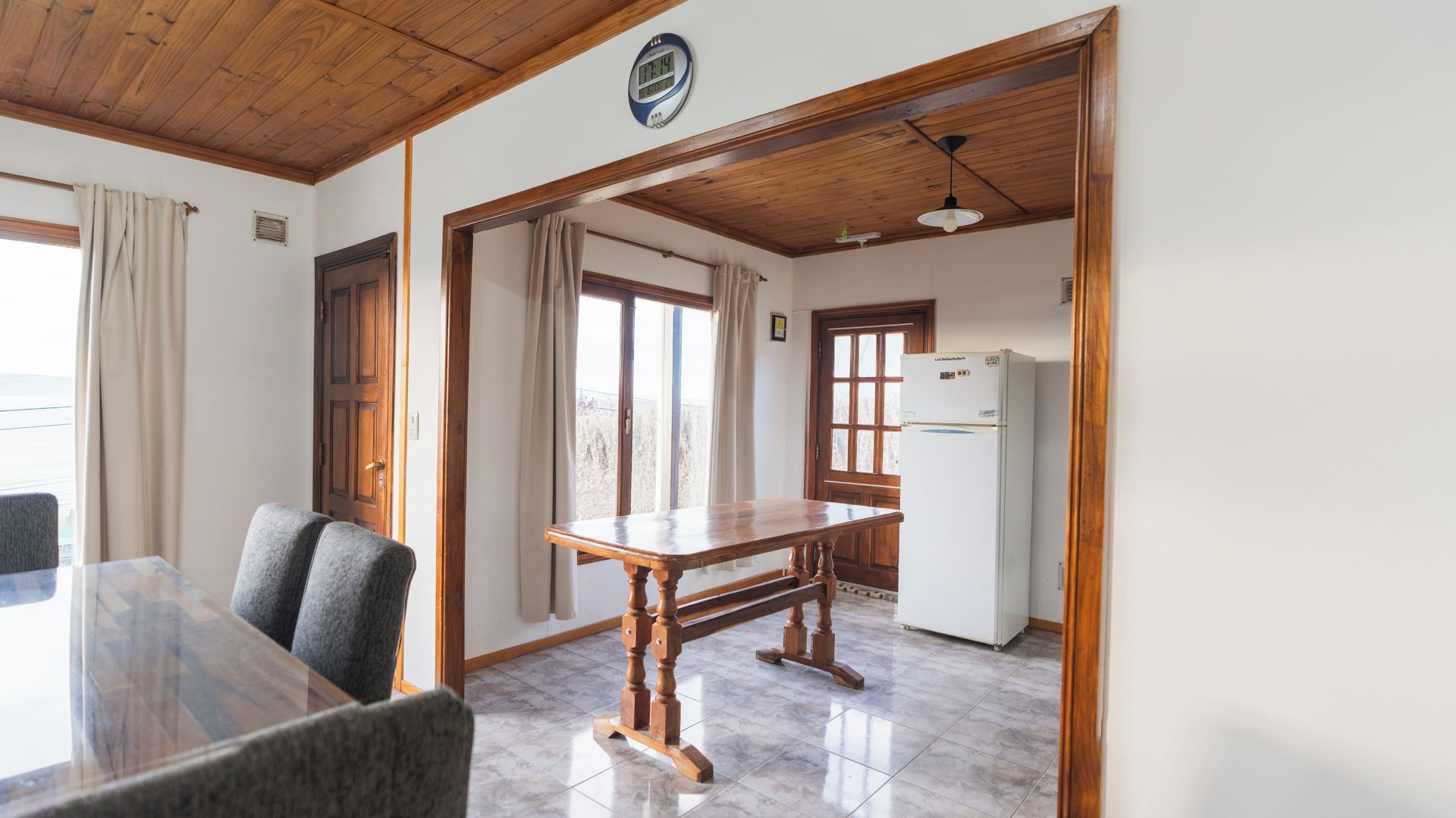 A room with a wooden ceiling, a dining table with chairs, a refrigerator, and a window with curtains.