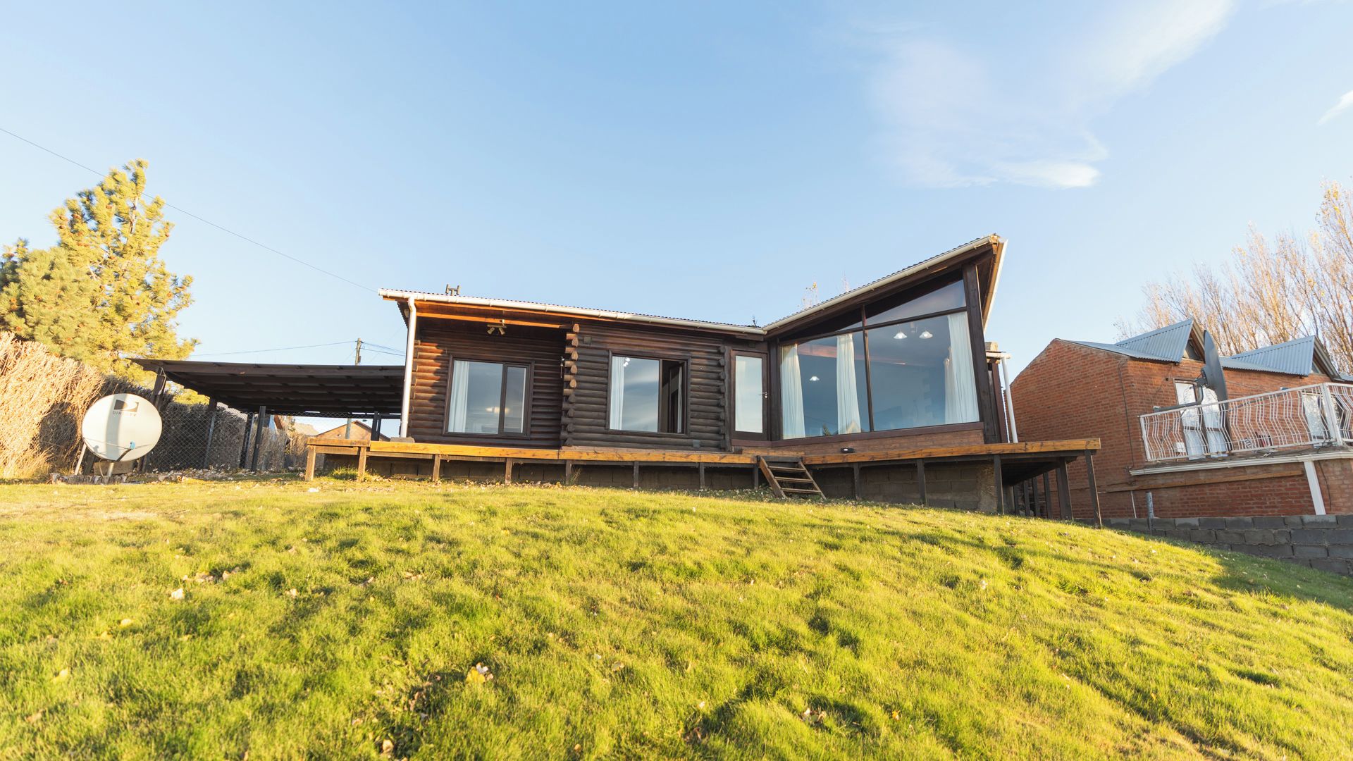 A wooden cabin with large windows, a deck, and a grassy hill.