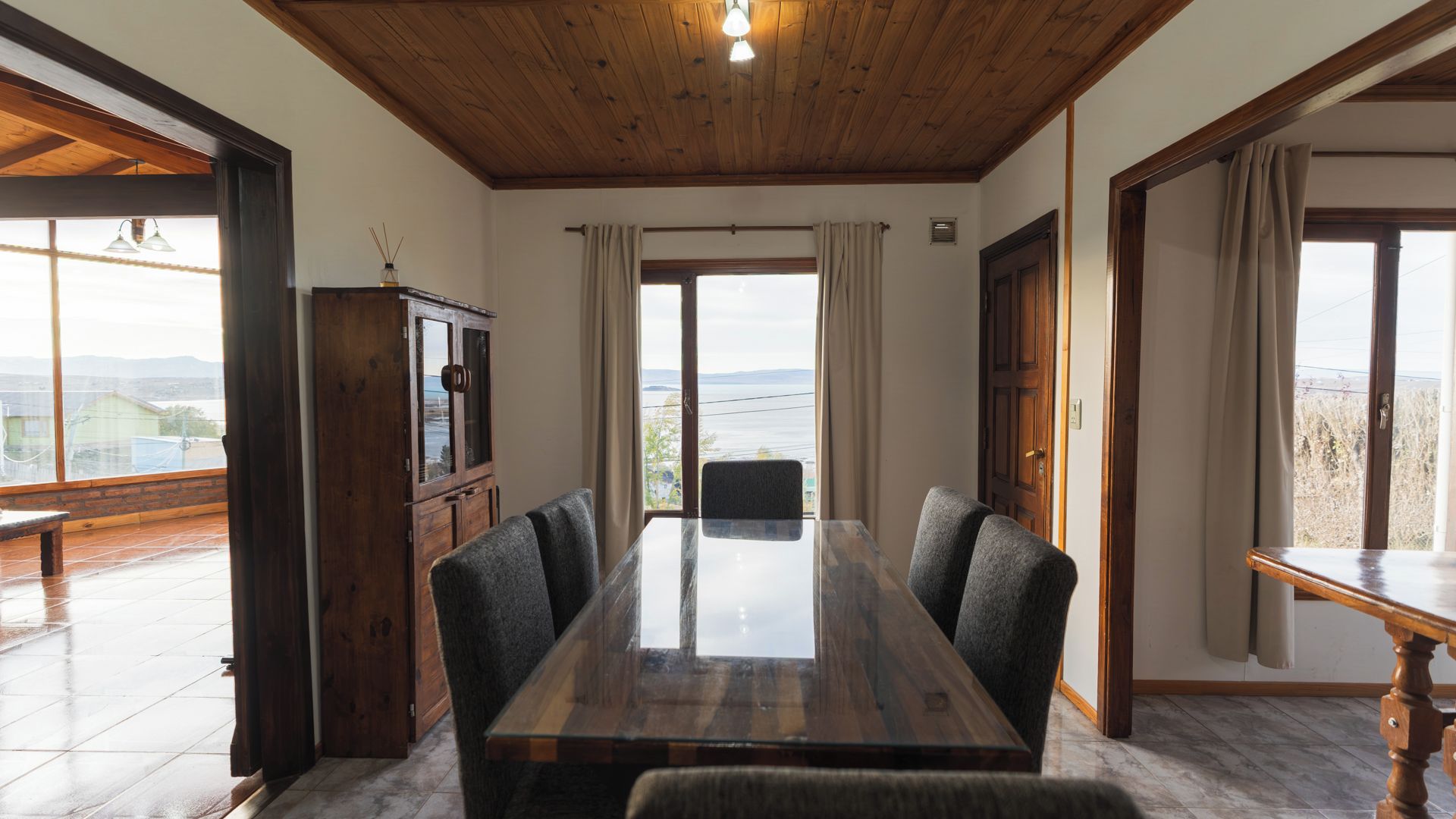 A dining area with a wooden table and chairs, a cabinet, and large windows with a view.