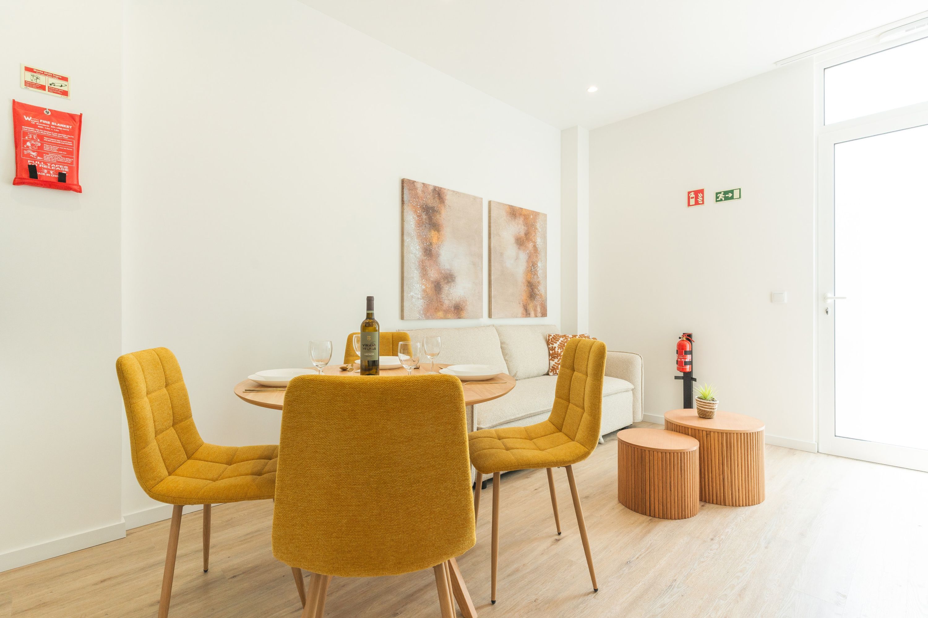 Un coin repas avec une table ronde, quatre chaises jaunes, deux petites tables d’appoint et une bouteille de vin.