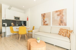 Ein modernes Wohnzimmer mit einer Couch, zwei Sesseln, einem kleinen Tisch und einer Küchenzeile inklusive Mikrowelle.