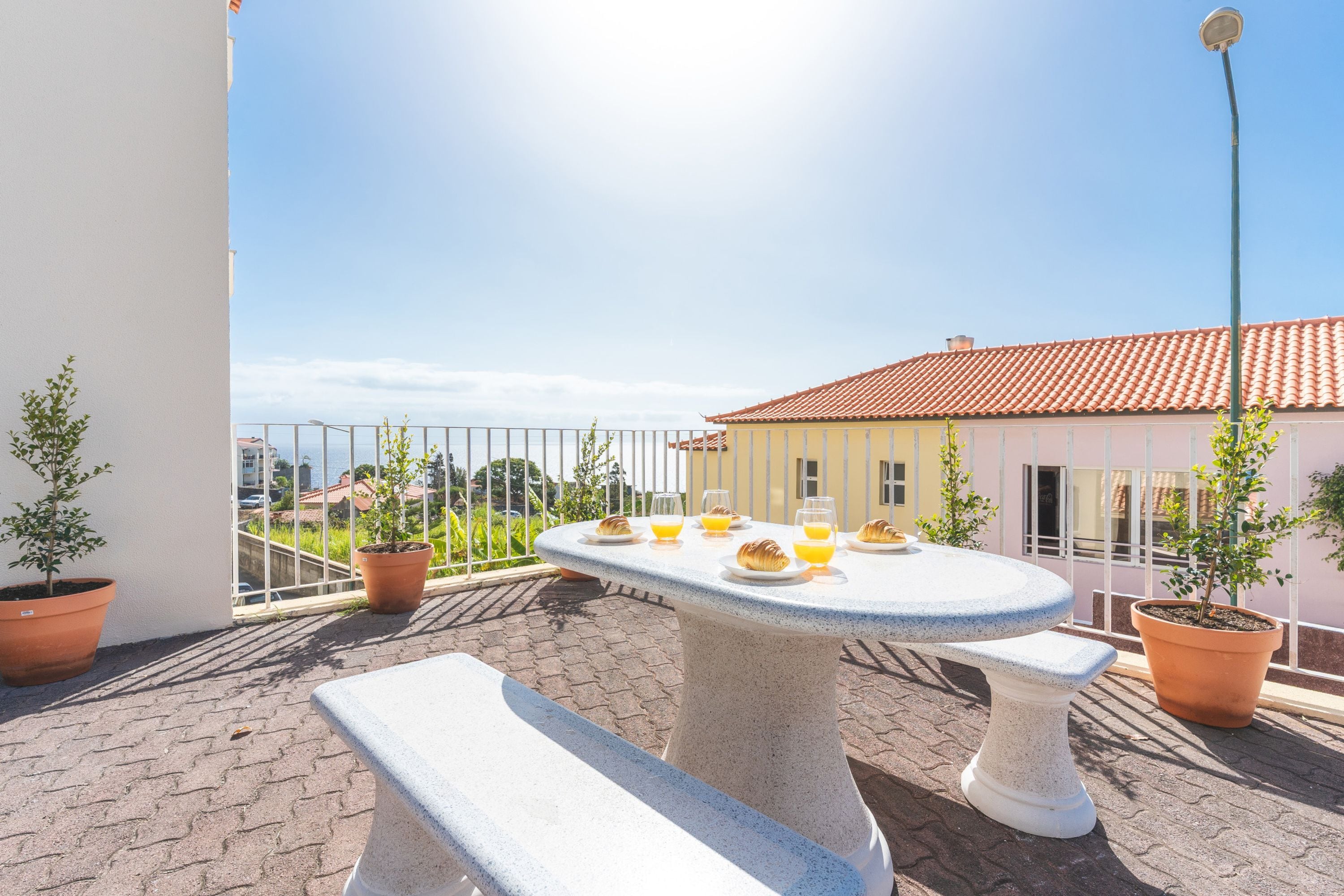 Una terraza con una mesa redonda dispuesta para el desayuno, plantas en macetas y vistas a un edificio con tejado de baldosas.