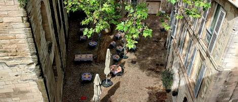 An outdoor seating area with tables and chairs, surrounded by stone walls and a tree providing shade.
