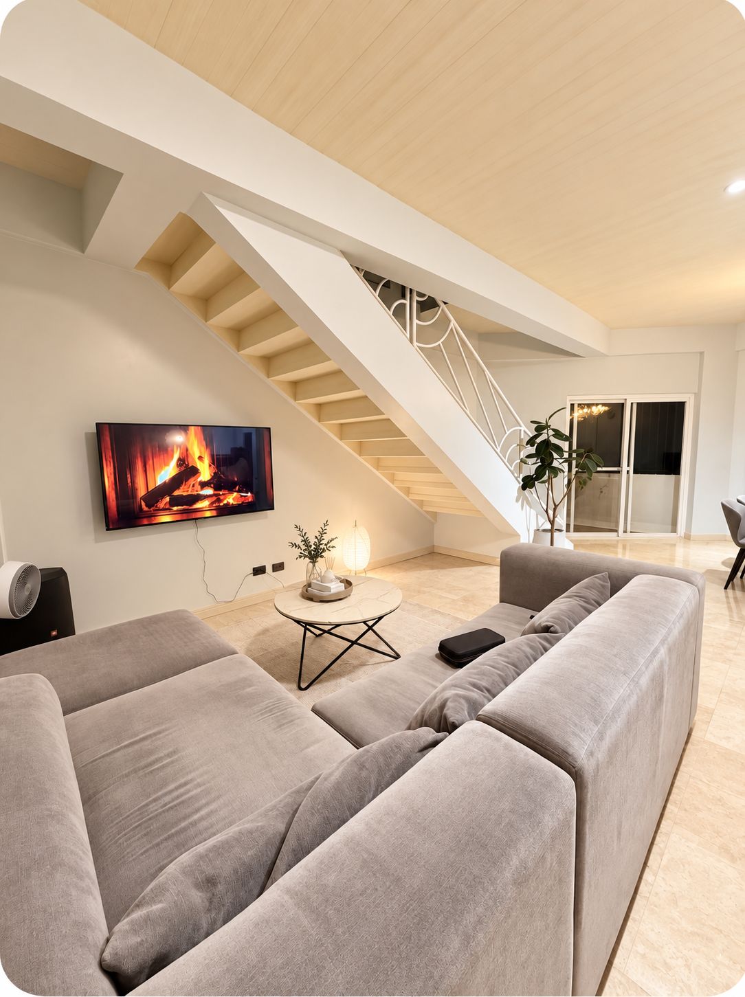 Een moderne woonkamer met een grote grijze bank, een flatscreen-tv waarop een open haard te zien is, een klein rond tafeltje met een plant en een trap naar een hoger gelegen verdieping.