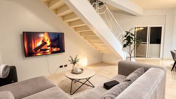 Een moderne woonkamer met een grote grijze bank, een flatscreen-tv waarop een open haard te zien is, een klein rond tafeltje met een plant en een trap naar een hoger gelegen verdieping.