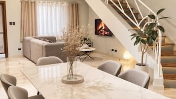 A modern living room with a marble dining table, grey upholstered chairs, a white staircase, and a fireplace.