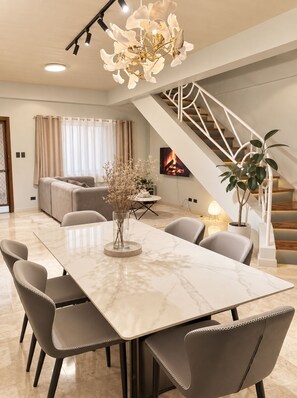 A modern living room with a marble dining table, grey upholstered chairs, a white staircase, and a fireplace. - Casa Alegre Spacious Home - Rooftop Tall View, Near Tourist Spots and Malls (Tagaytay City)