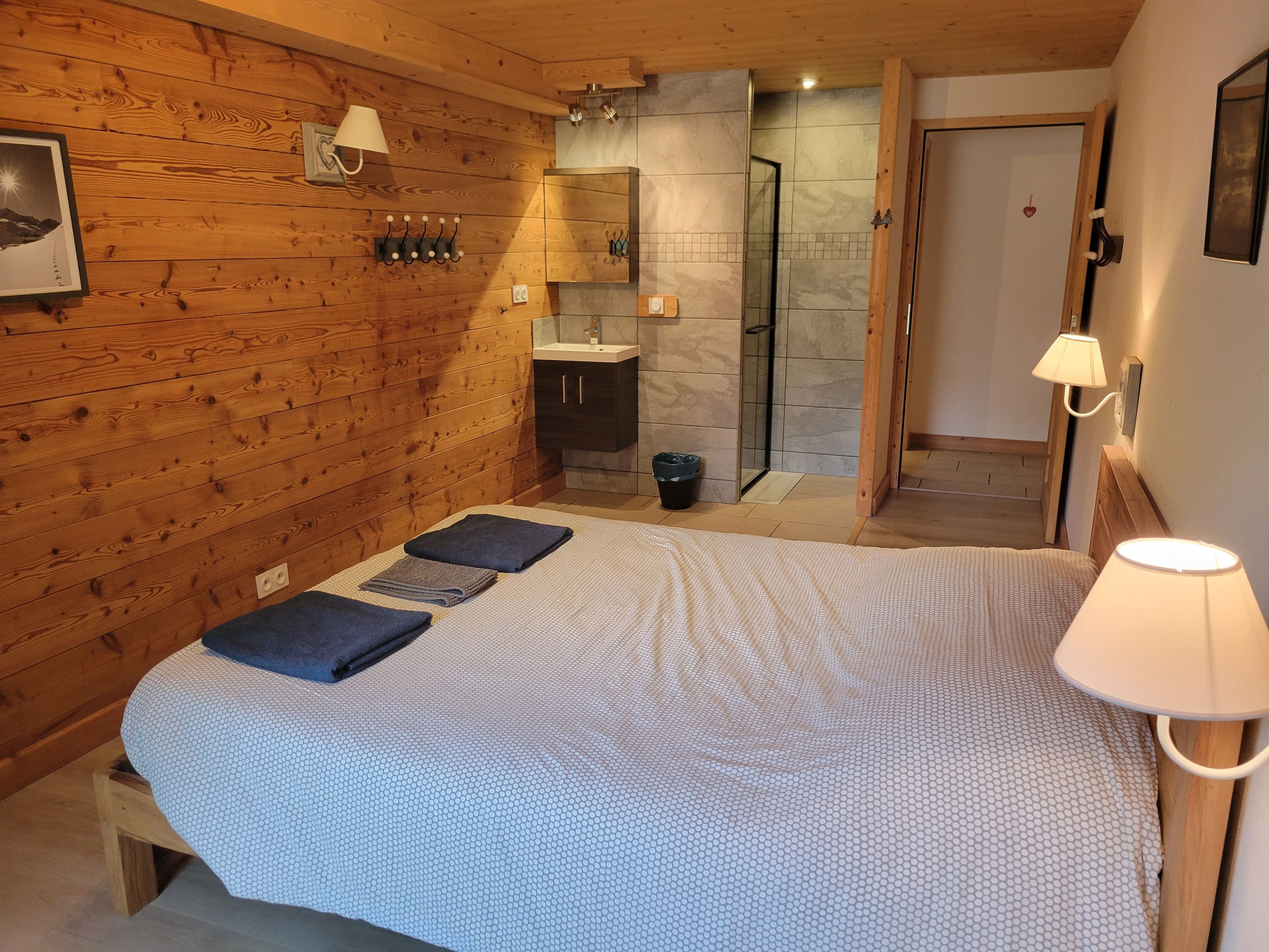 Une chambre à coucher avec un lit, une table de chevet, une lampe, un mur en bois, une salle de bain avec une douche et un miroir.