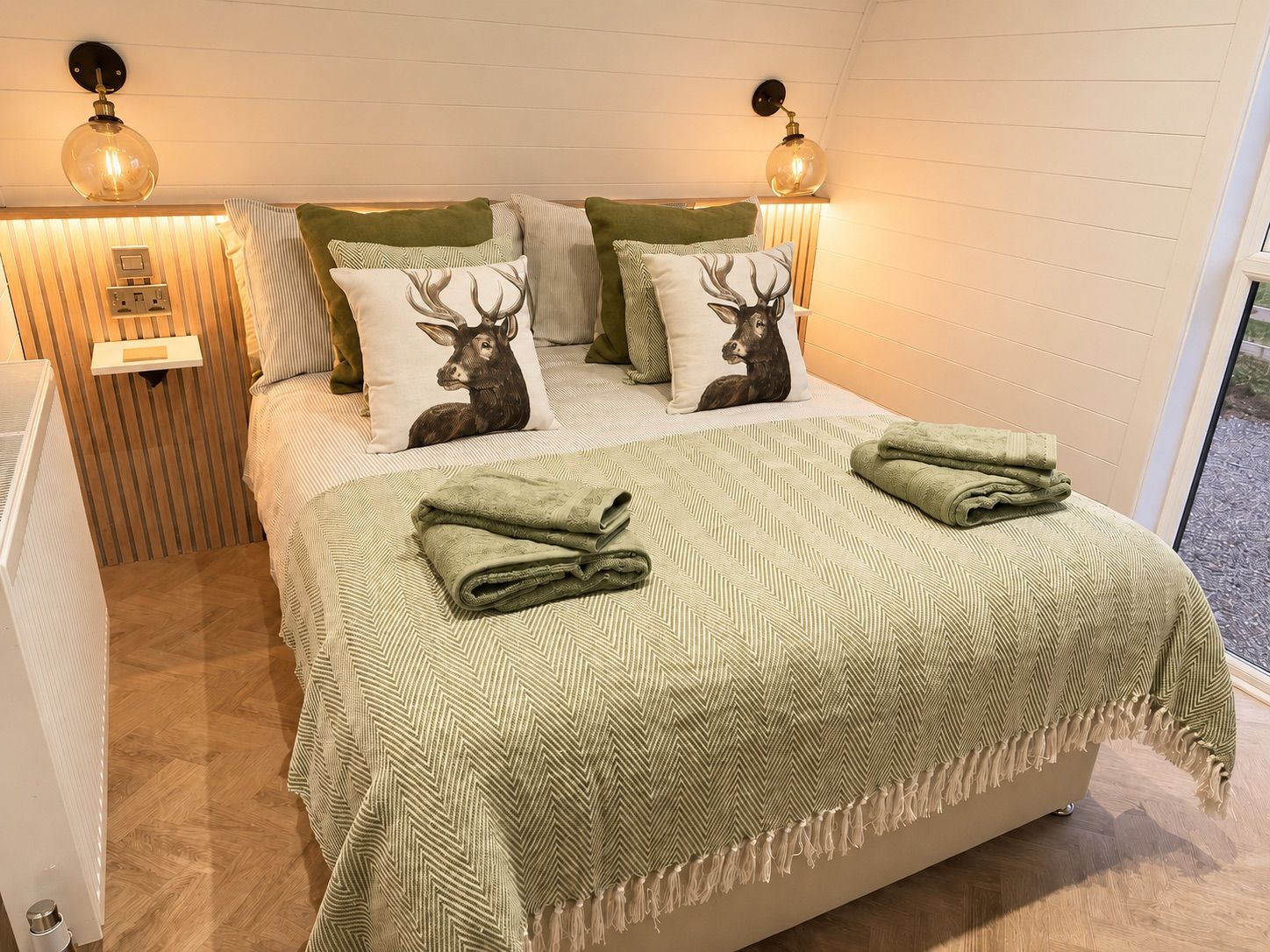 Una cama con una colcha y almohadas estampadas, una mesita de noche con una lámpara, y vistas al exterior a través de una ventana.