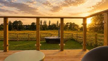 Eine Holzterrasse mit Blick auf ein grasbewachsenes Feld und Bäume, ein Whirlpool und ein überdachter Sitzbereich.