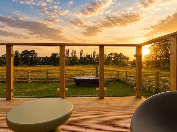 Una terraza de madera con vistas a un campo cubierto de césped y árboles, un jacuzzi y una zona de estar cubierta.