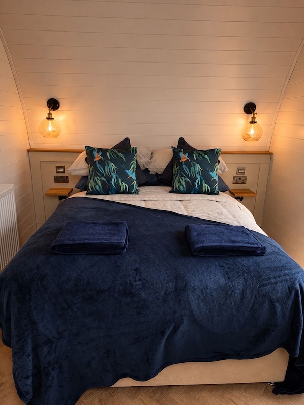 A bed with a dark blue cover, two pillows with a tropical print, and two bedside lamps.