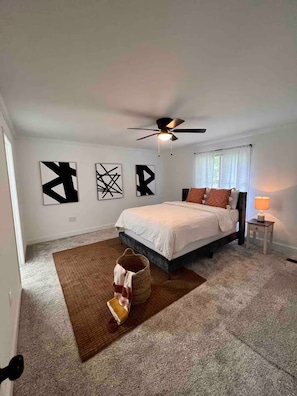 Ein Schlafzimmer mit Bett, Deckenventilator, Teppich und abstrakten Wandbildern.
