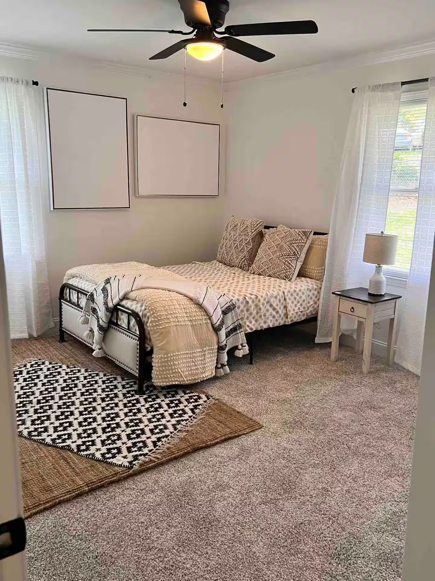 A bedroom with a bed, a ceiling fan, a nightstand, a lamp, a rug, and a window with curtains.