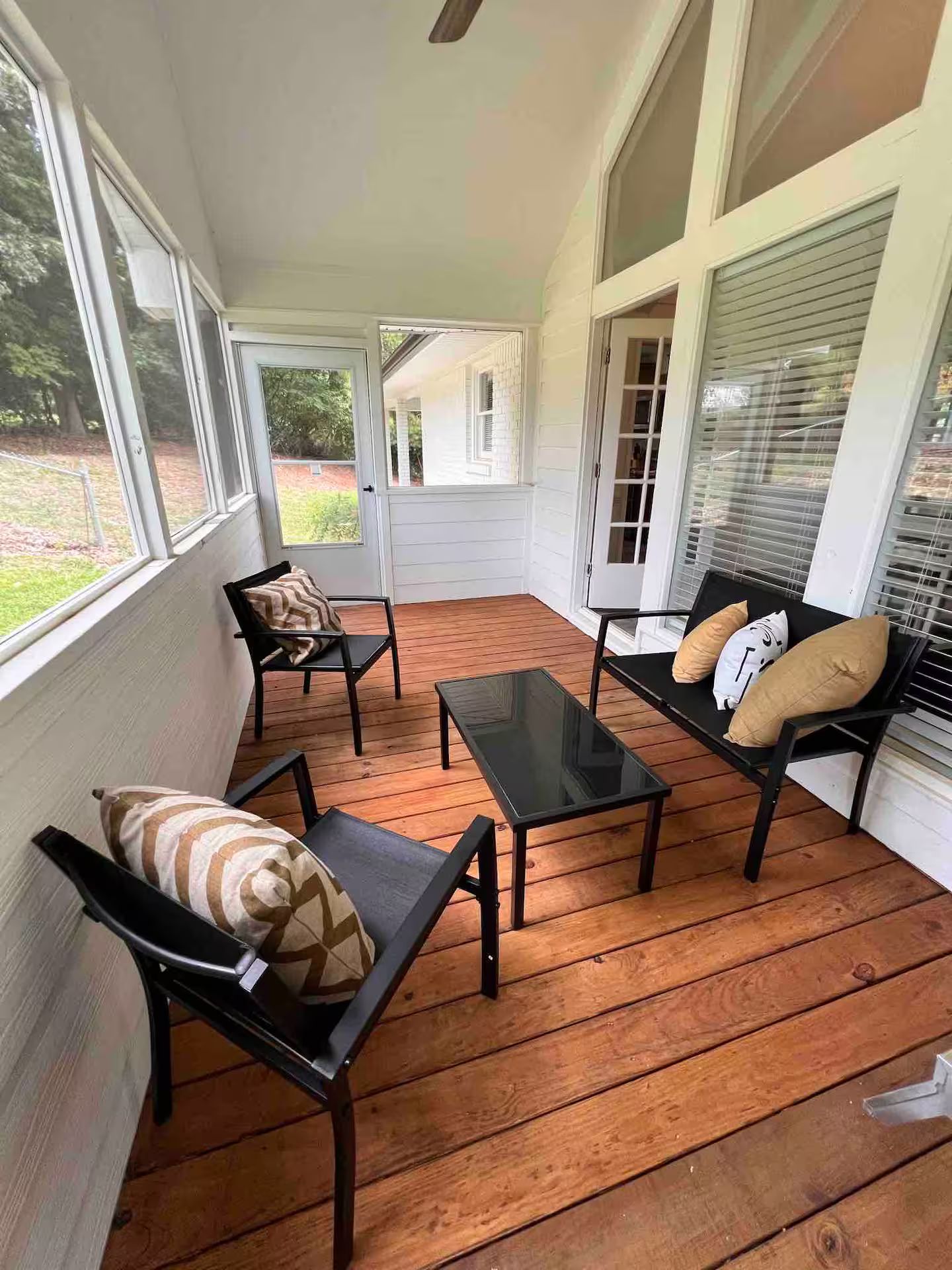 A covered porch with wooden flooring, black metal furniture, and large windows.