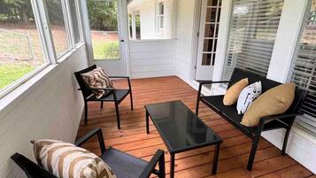 A covered porch with wooden flooring, black metal furniture, and large windows.